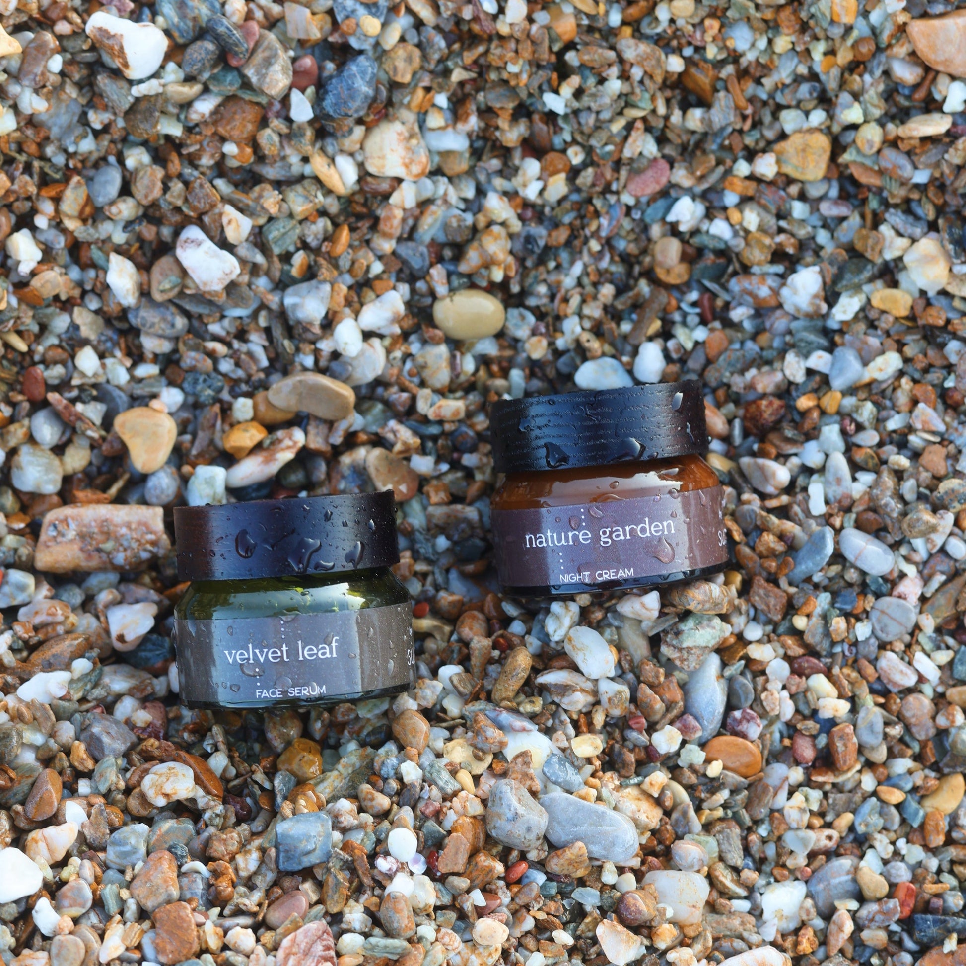 Conscious herbal skincare jars labeled 'velvet leaf' and 'nature garden' on a pebbly beach.