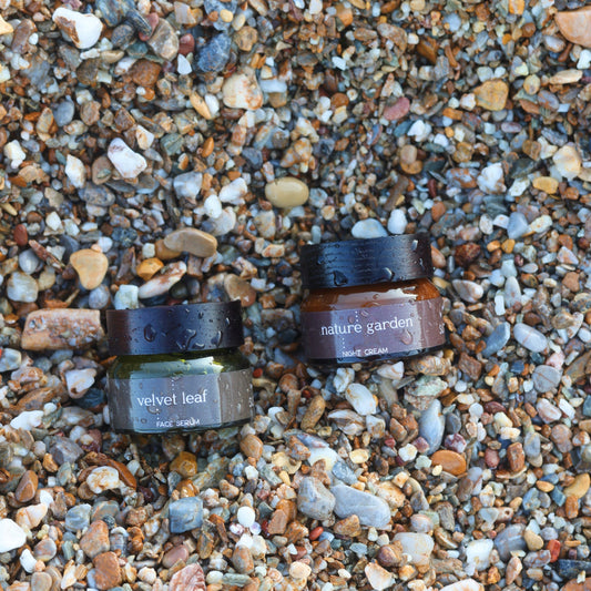 Conscious herbal skincare jars labeled 'velvet leaf' and 'nature garden' on a pebbly beach.